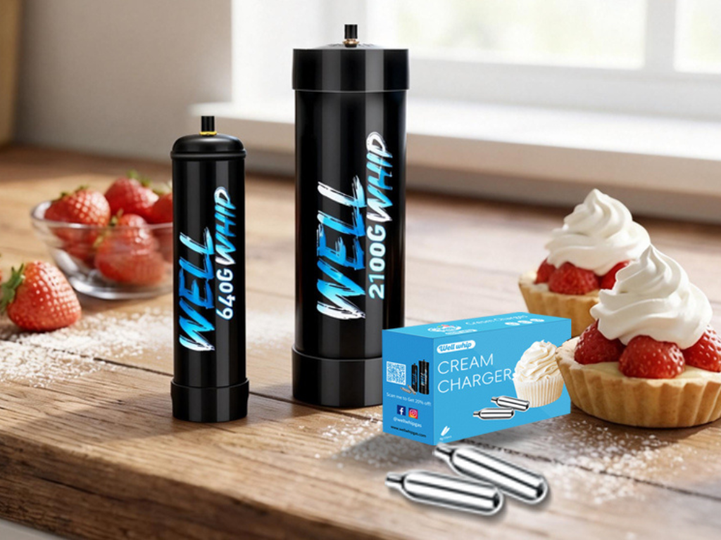 Two black cream chargers labeled "WELL 640G WHIP" and "WELL 2100G WHIP" on a wooden counter with fresh strawberries and whipped cream tarts nearby.