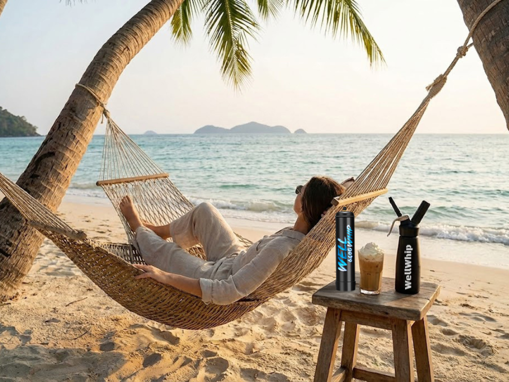 A person relaxes in a hammock between palm trees on a tropical beach. Nearby, a table holds a drink topped with whipped cream and two whipped cream chargers. The scene feels serene and peaceful.