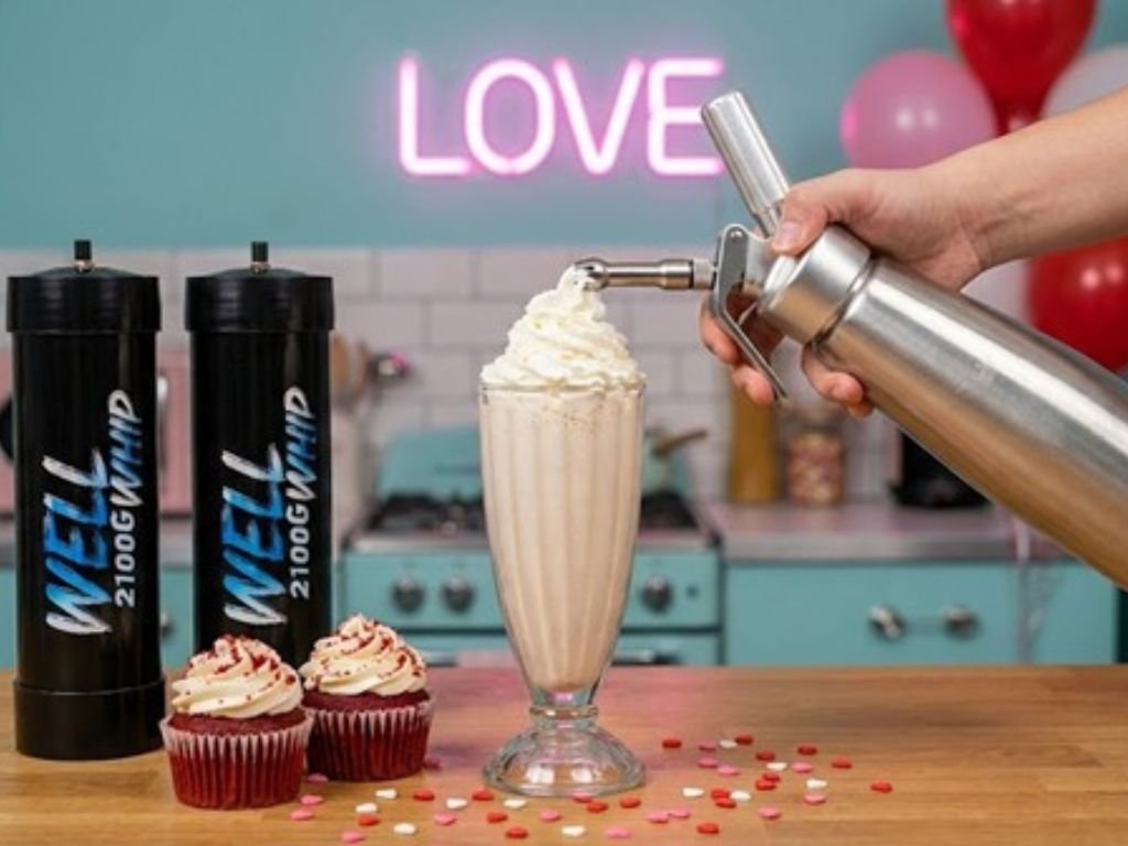 A hand tops a milkshake with whipped cream using a dispenser. Nearby are two red cupcakes. A neon sign reading "LOVE" glows in a retro-styled kitchen.