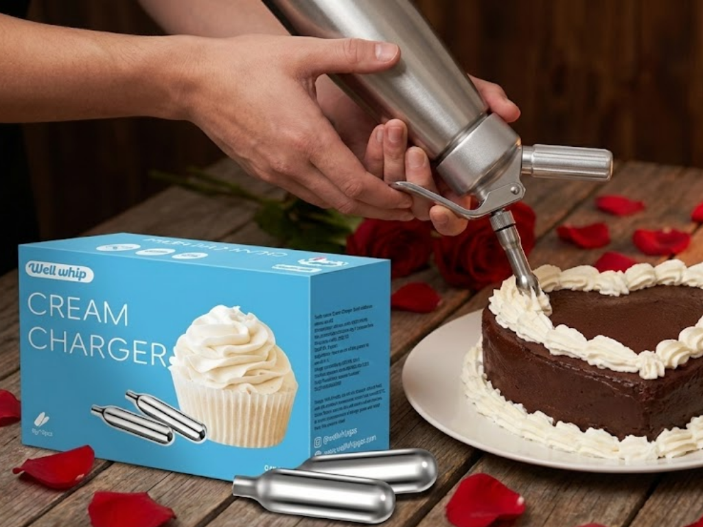 A person decorates a heart-shaped chocolate cake with whipped cream using a dispenser. A Well Whip cream charger box and rose petals are visible.